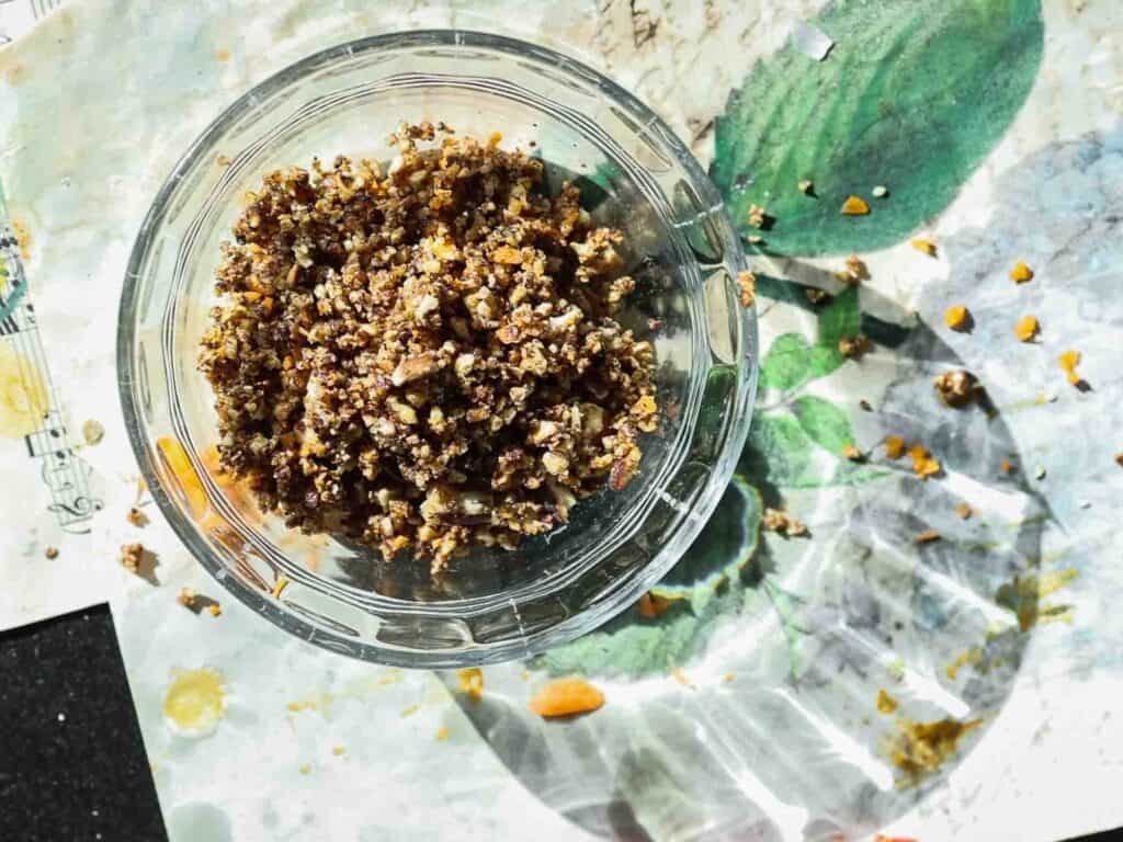 A clear glass bowl filled with a mixture of chopped nuts on a patterned surface. Some crumbs are scattered around the bowl, catching the sunlight.